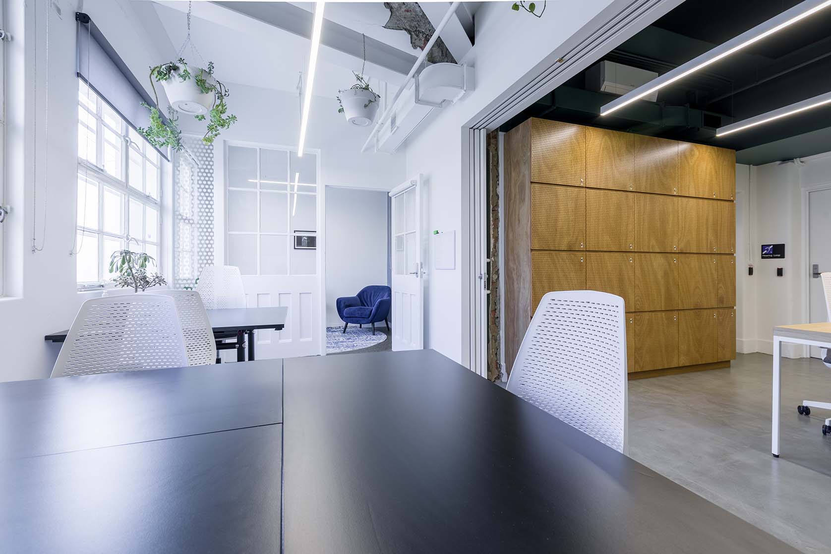 Brightly lit co-work space with black desks, white chairs and wooden lockers on the right..