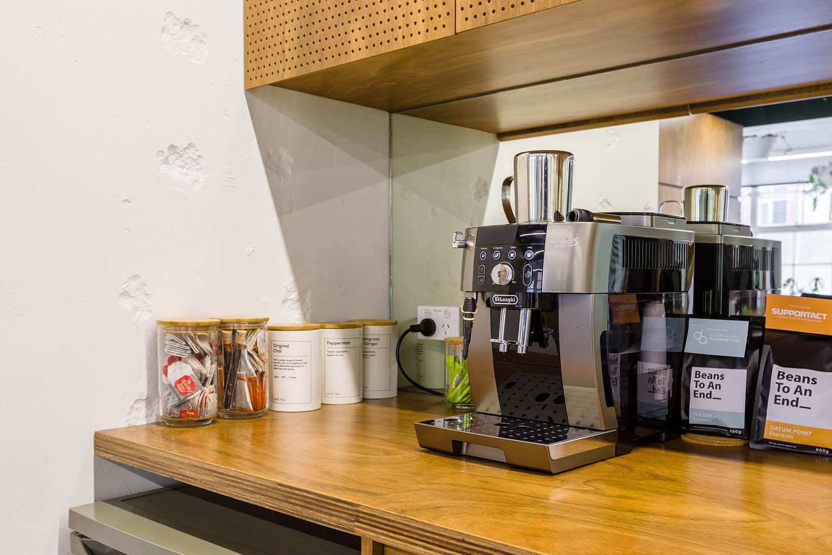 Photo of silver and black coffee machine in the corner of a wooden kitchenette. To the left are four ceramic canisters of tea and on top of the coffee machine is a silver jug.