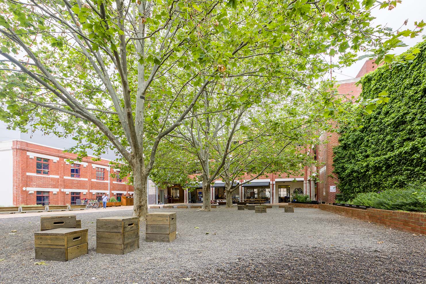 Photo of four wooden crate seats are scattered in the bottom left on grey gravel flooring. To the right is a large tree with green foliage, and in the background is a red brick building.