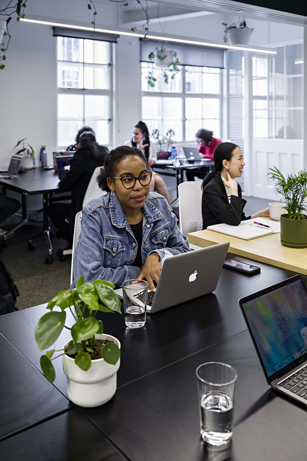 Photo of woman sitting at a desk in a co-work space, with other workers in the background surrounding her. On the desk is her laptop, phone, glass of water and a plant in a white pot. The room is brightly lit and other workers are talking in the background.