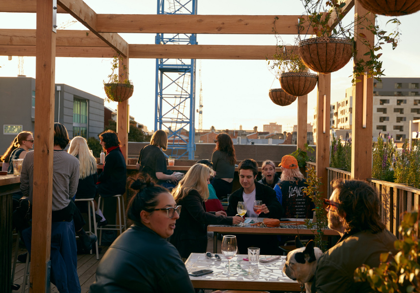 Photo of people socialising at Runner Up Rooftop Bar. In afternoon sun the people are sitting at mosaic tables, the space framed by wooden rafters and hanging plants.