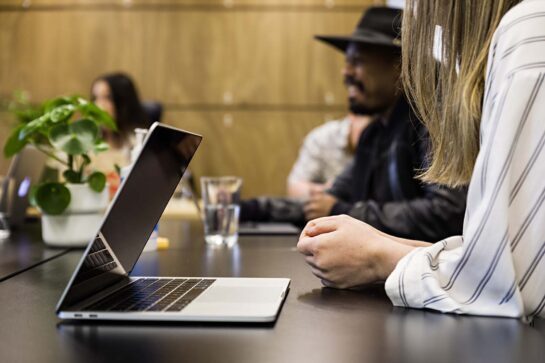 Photo of laptop on a desk in a coworking space, with people working and talking in the background.