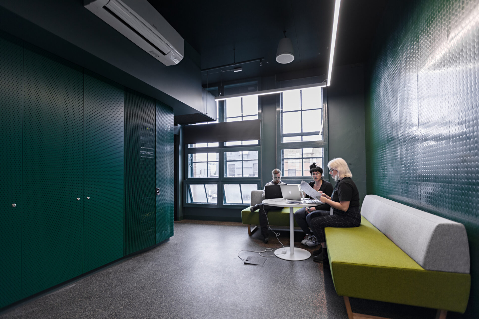 Dark meeting room with dark green walls and bright green and grey couch to the right. Three people are meeting at a table in the corner using laptops and paper, with large window panels behind them.