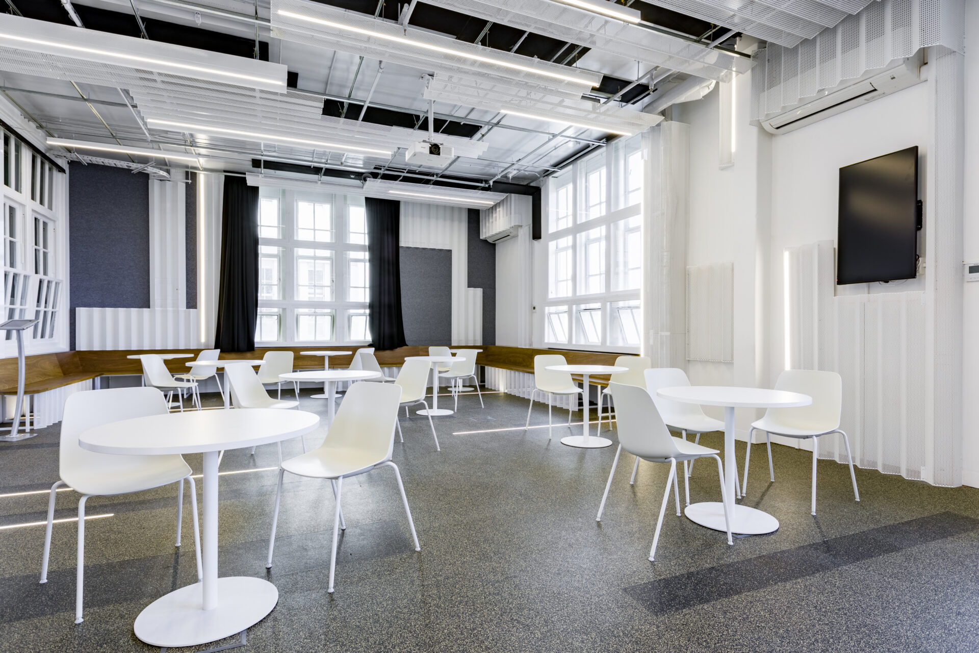 Event space at music market filled with white tables and chairs. The flooring is grey, and the walls are white and grey. The room is lit with bright natural light from large panelled windows.