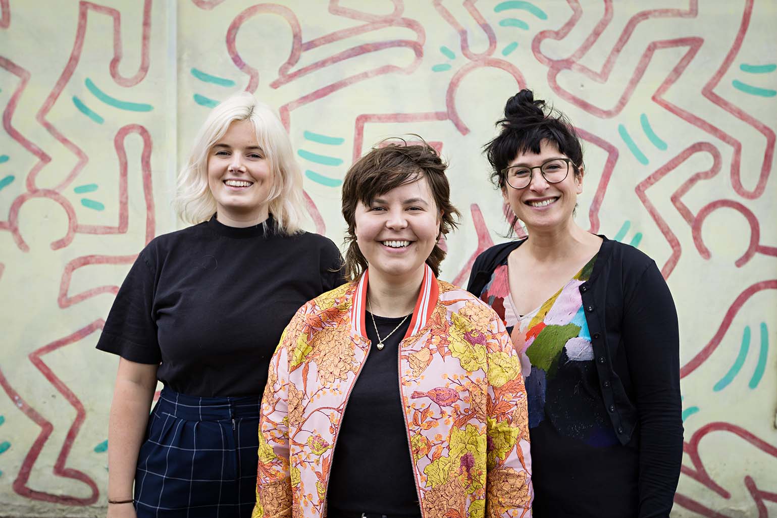 Photo of three people smiling at the camera in front of a white, red and green mural.