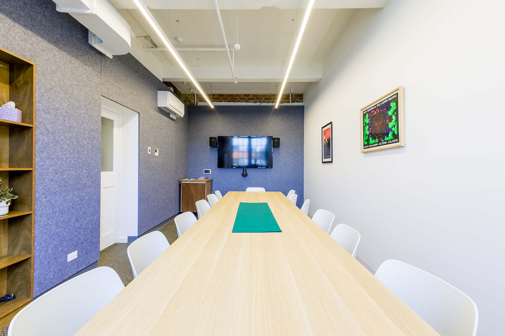 Photo of board room. At the centre is a long wooden table with white chairs leading towards the back of the room. The left and back walls are blue, and the right wall is white. There is a white door to the left, a tv on the back wall, and two art prints on the right.