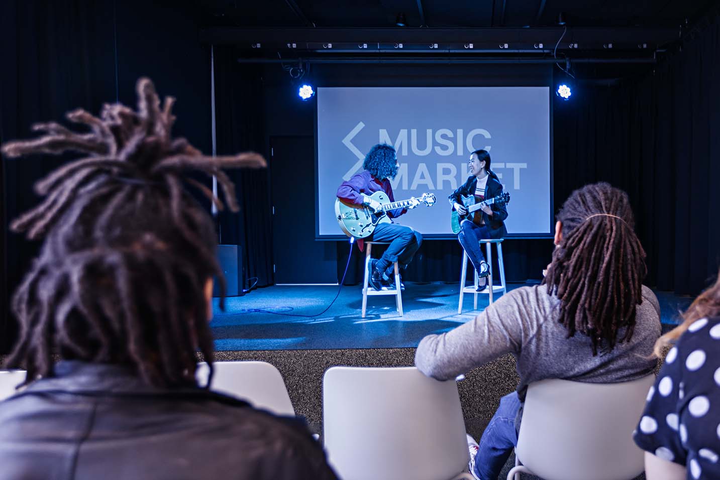 Photo from the perspective of the crowd in Music Market event space. In the centre is a blue-lit stage with two people playing guitar on stools. Behind them is a projector screen that says Music Market in white writing.