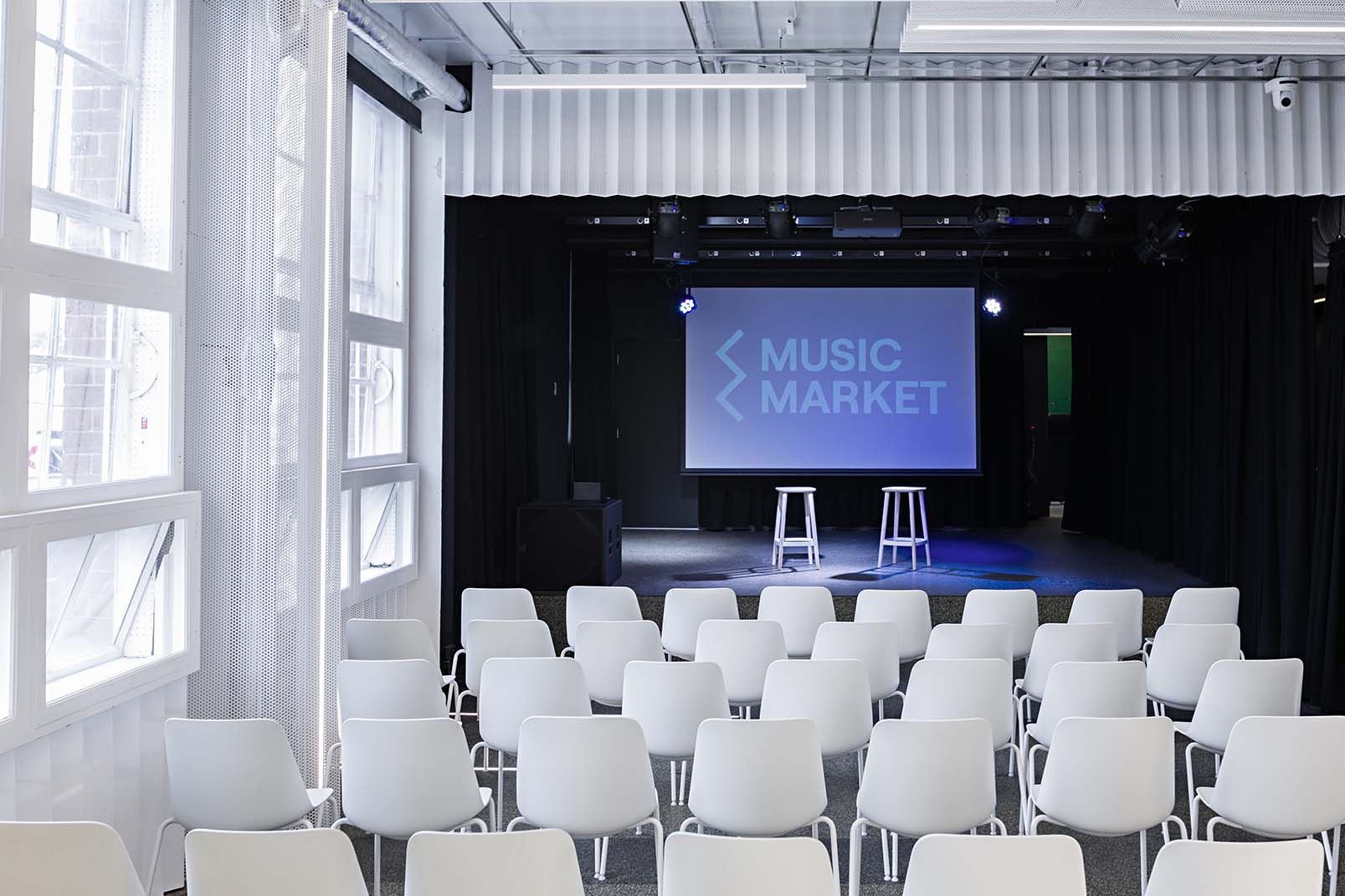 Indoor event space with chairs set up for seated audience facing stage. Stage is set up with two stools and projector screen that says Music Market.