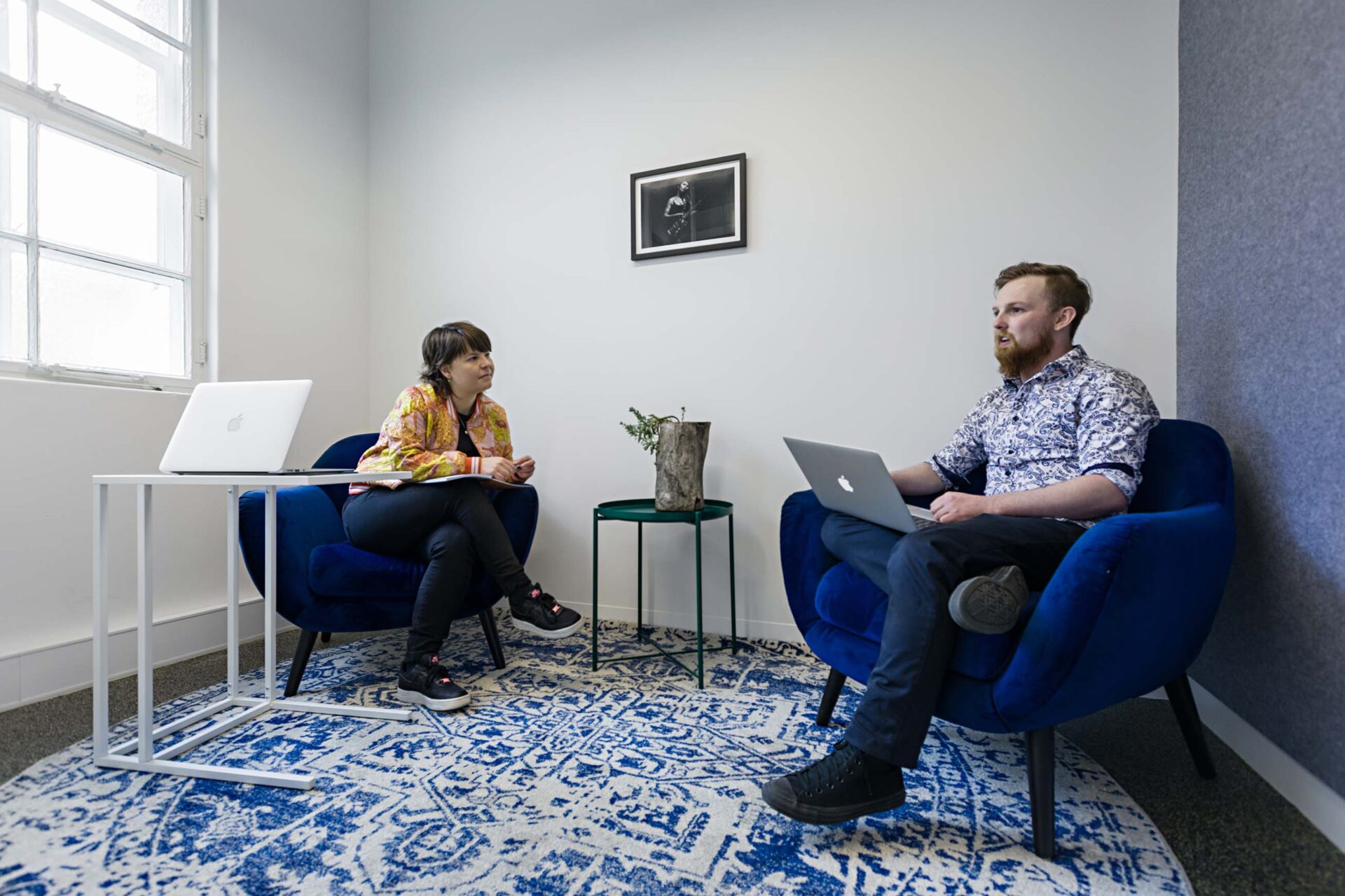 Two people meeting in co-work space, sitting on blue chairs and using laptops. The rug below them is blue and white.