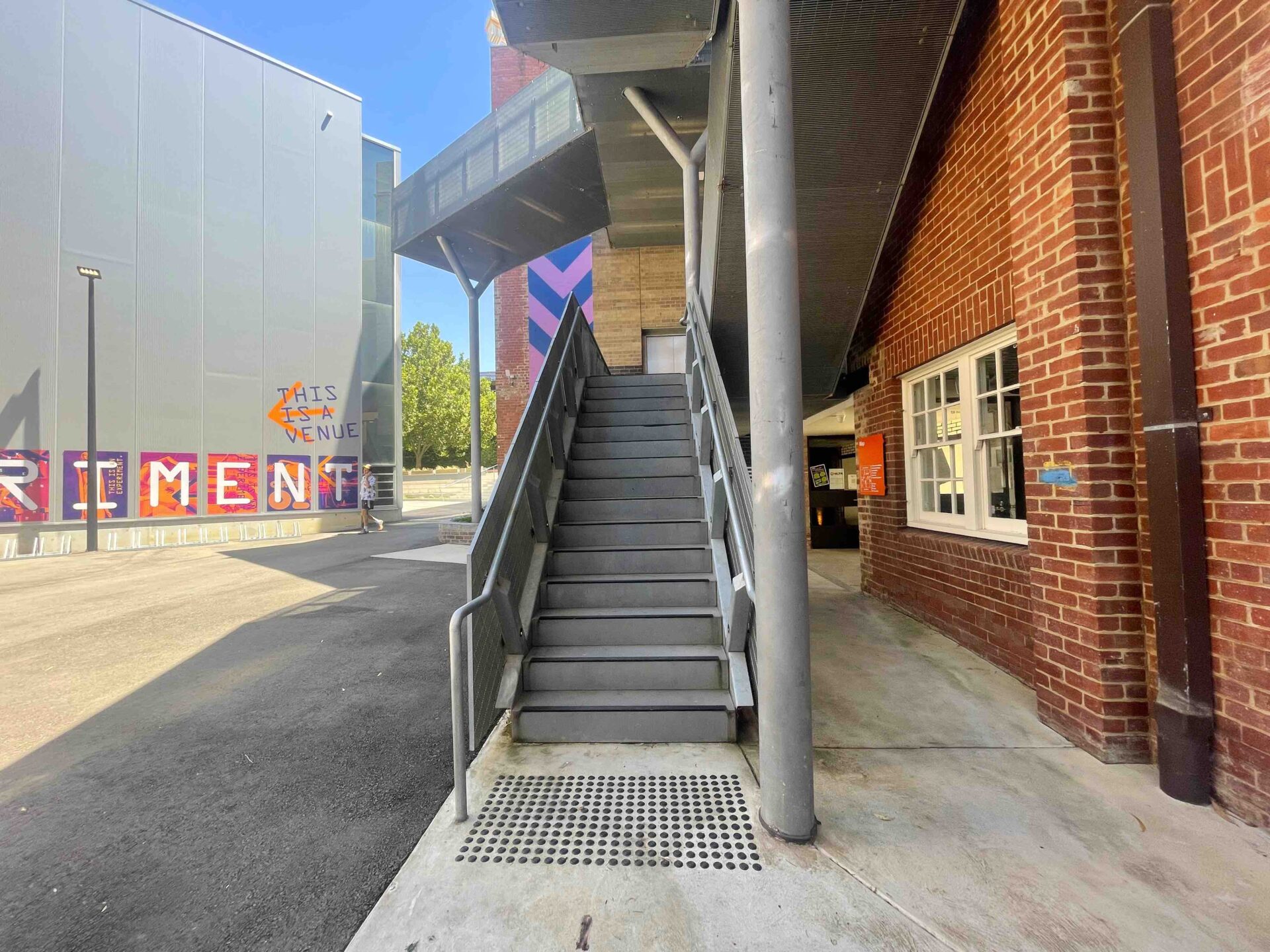 Photo from the bottom of an outdoor concrete staircase looking directly up the stairs. To the right is a red brick building with white windows, and to the left is a tall grey-panelled building with bright pink and purple posters on each panel. Above the staircase are some metal landing spaces that the stairs lead to.