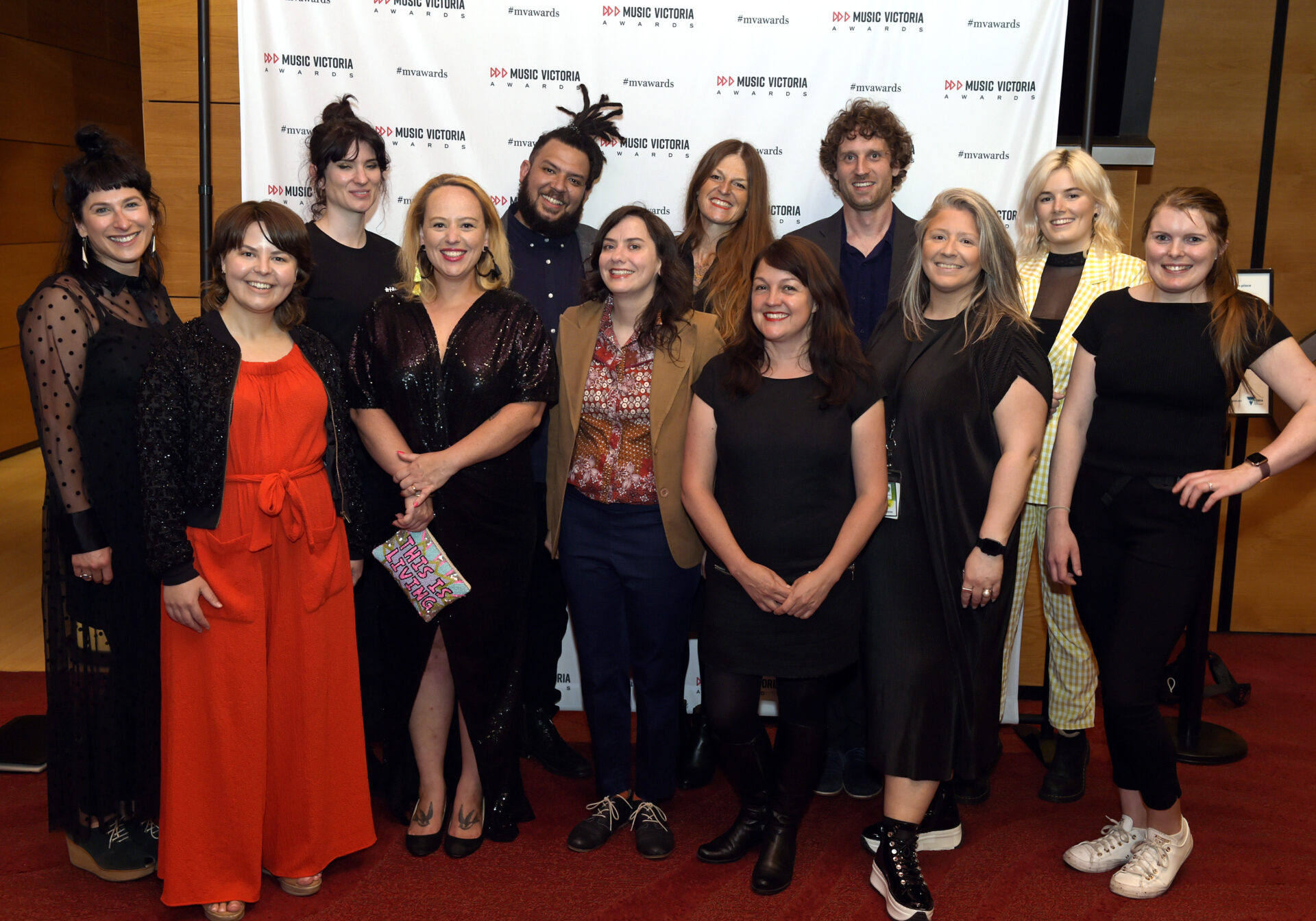 Photo of twelve Music Victoria staff members standing in front of a white banner with a Music Victoria Awards logo. They are dressed for the event and are standing on a red carpet.