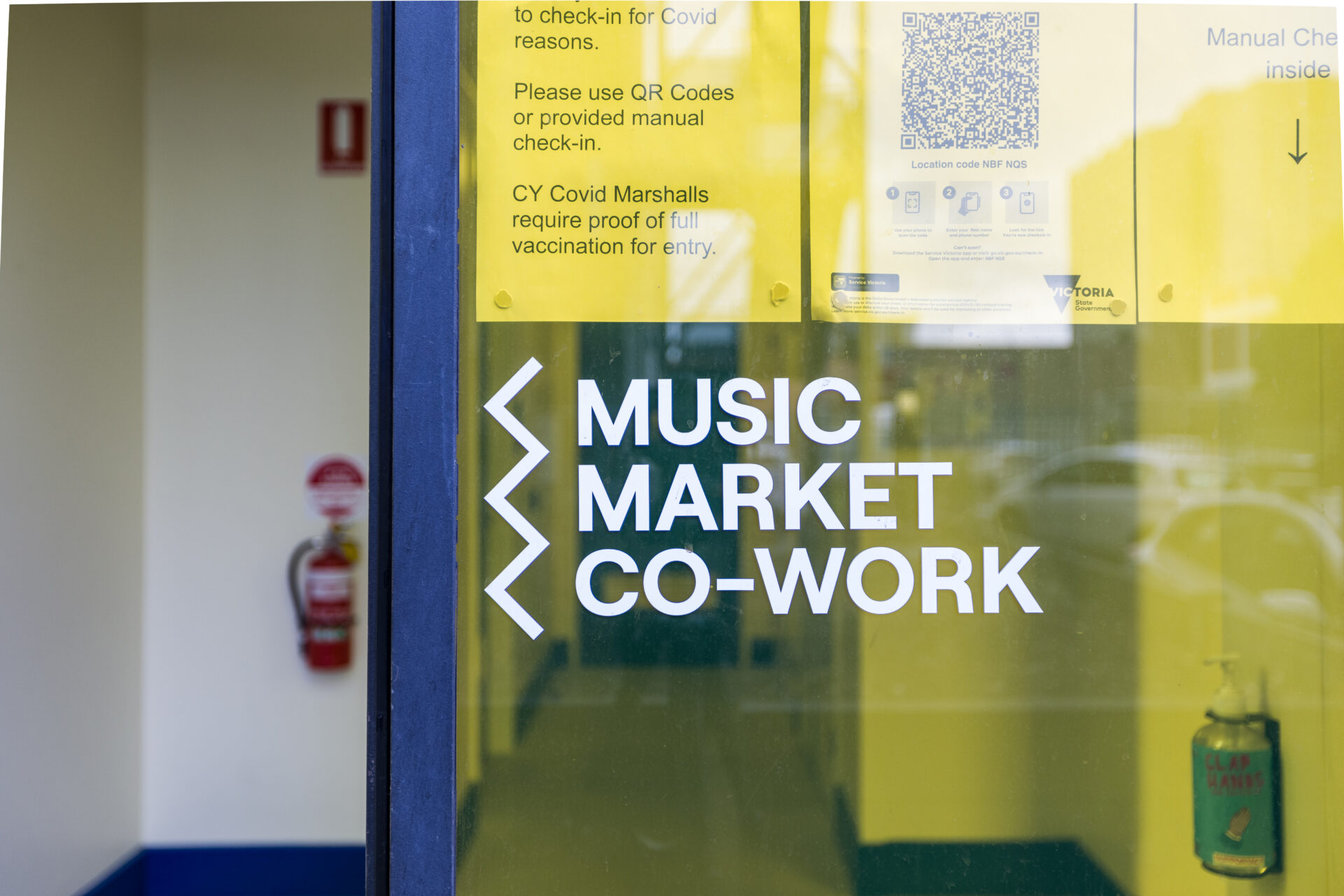 Photo of a yellow glass window with an open doorway to the left. On the window is a white logo that has a zig zag line to the left and the words "Music Market Cowork" in bold uppercase letters. Above that are Covid check-in print outs and guidelines.