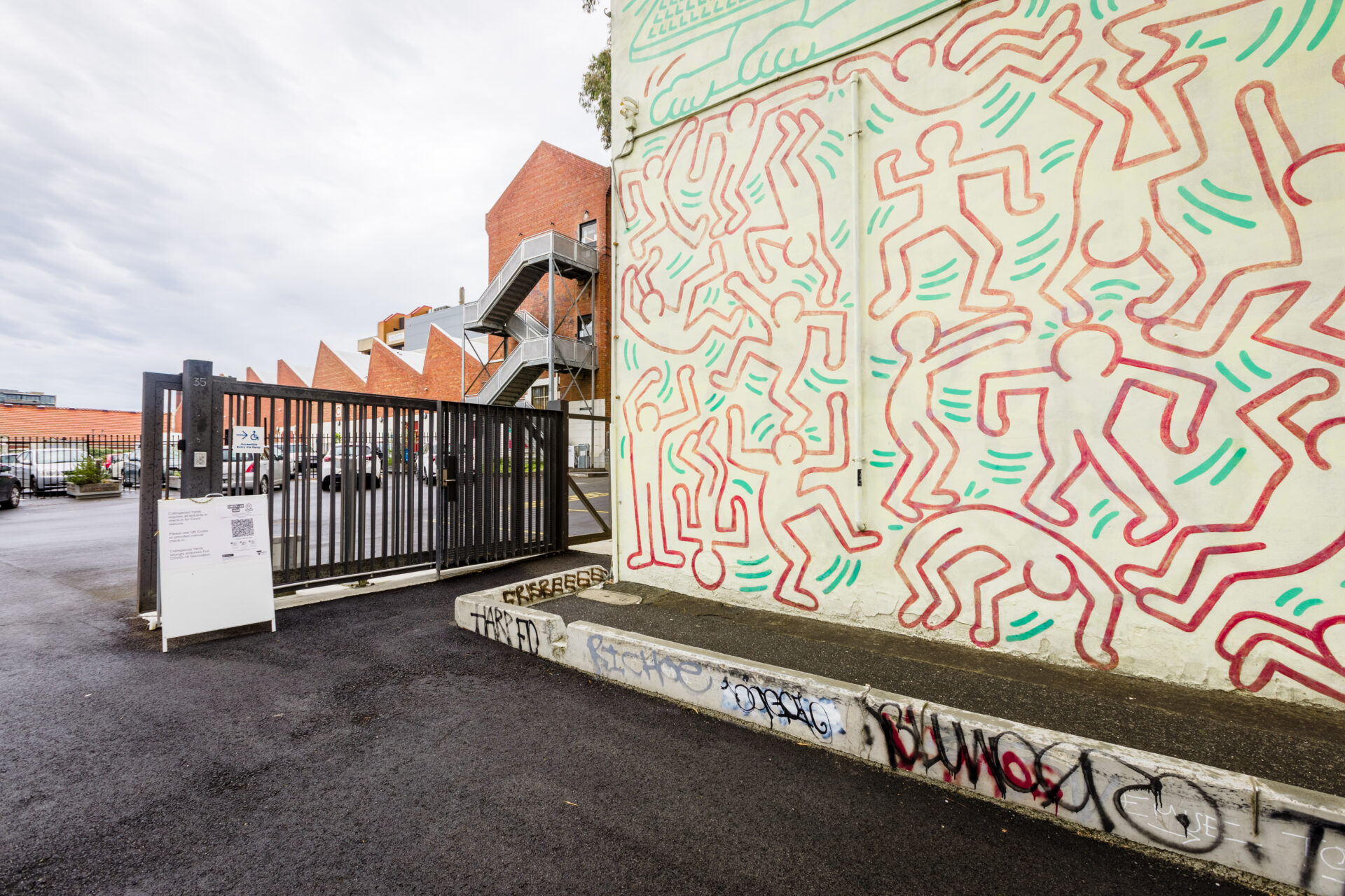 Photo of Collingwood Yards Keith Haring mural. The mural is painted outlines of people characters with scattered green lines in between. To The left of the mural is a security gate. The ground is black asphalt and there is some graffitied concrete below the mural.