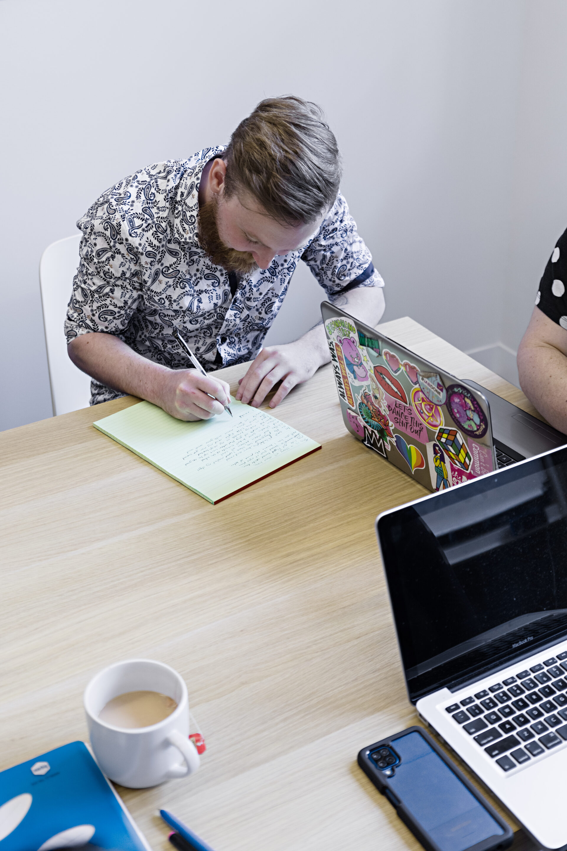 Photo of two people working at a wooden desk, one to the left and one is cropped out of the photo on the right. The first person is hand-writing on a yellow note pad and wearing a black and white patterned shirt. The person on the right is working at a grey laptop covered in brightly coloured stickers.