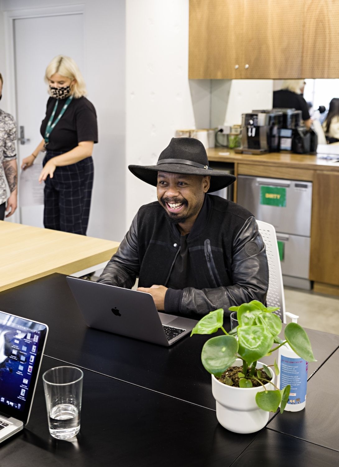 Photo of person working at a laptop in a co-work space, smiling across at another worker. There is a plant on the table. In the background to the left there are people standing, and to the right there is a kitchenette.
