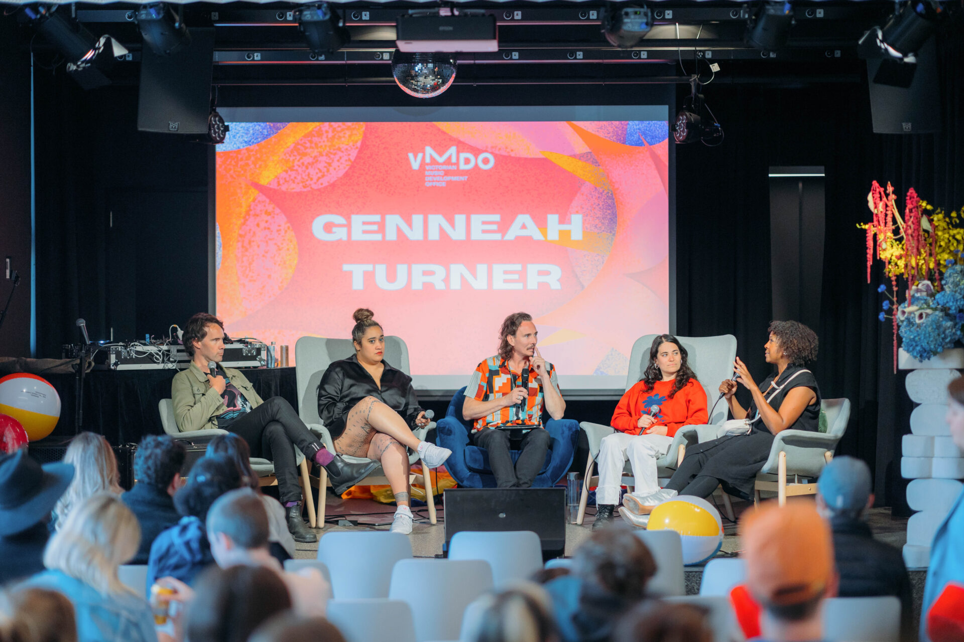 Photo of five people sitting on grey seating on an indoor stage speaking to each other with microphones. A crowd is watching the stage. In the top middle there is an orange, pink and blue projection on a screen, with bold, uppercase white writing that says Genneah Turner. Above this is the VMDO logo.
